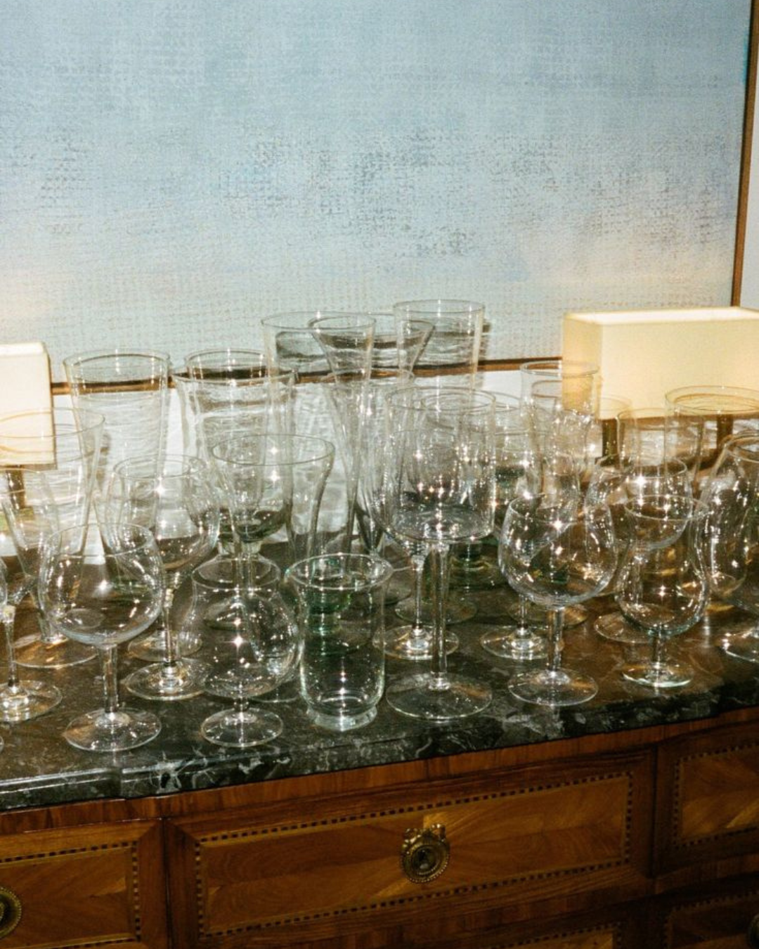 Collection de verres en cristal vintage soigneusement disposés sur un buffet en bois avec un fond bleu texturé - clarisse demory