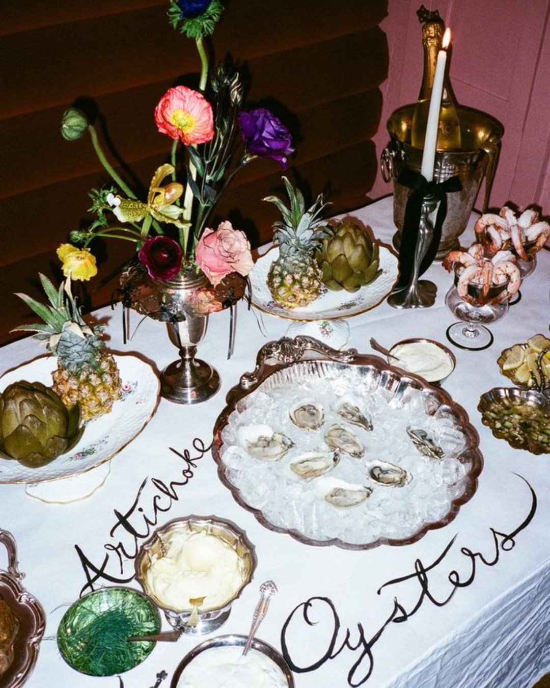 Table festive avec des fleurs colorées, des fruits exotiques, des huîtres sur glace et des crevettes, avec un chandelier et une bouteille de champagne en arrière-plan