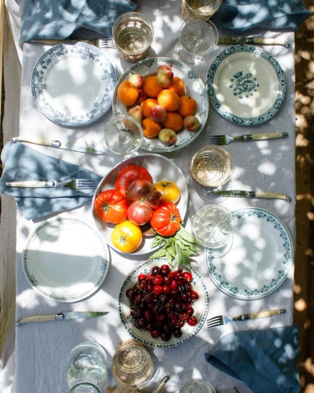 Table extérieure sous la lumière du soleil, dressée avec des assiettes en porcelaine bleue et un assortiment de fruits frais (abricots, tomates, cerises)